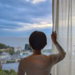 窓辺に立ち、カーテン越しに外を見つめる人物の後ろ姿